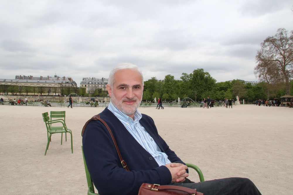 Elderly man with grey beard sitting on a green chair in a public park.